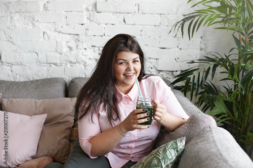 Horizontal shot of charming obese young female choosing healthy lifestyle drinking green spinach smoothie in modern living room, smiling happily, enjoying fresh taste. Weight loss and detoxication
