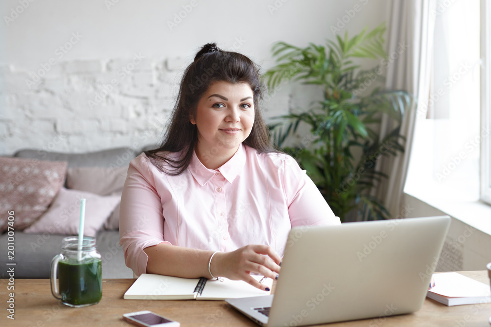 Cute body positive young chubby female freelancer sitting in modern room interior in front of open laptop with blank screen phone, glass of smoothie, copybook and decorative plant pot on table Cute body positive young chubby female freelancer sitting in modern room interior in front of open laptop with blank screen phone, glass of smoothie, copybook and decorative plant pot on table