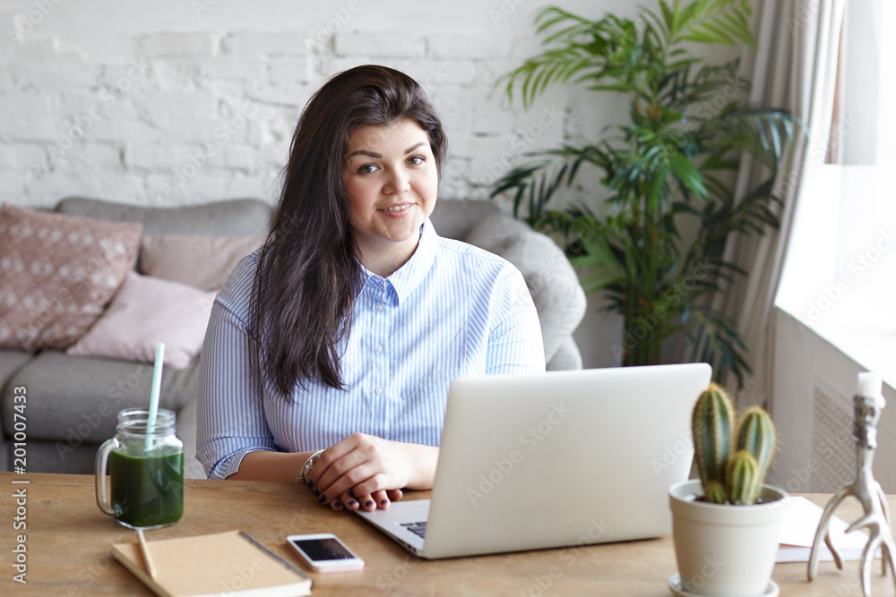 Picture of confident friendly looking young businesswoman with curvy body working on laptop pc at home office, sitting at her workplace, wearing stylish blue shirt, smiling broadly at camera Picture of confident friendly looking young businesswoman with curvy body working on laptop pc at home office, sitting at her workplace, wearing stylish blue shirt, smiling broadly at camera