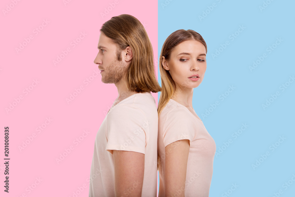 Curious creature. Beautiful young woman peeking into her boyfriends direction while standing back-to-back with him, posing against a blue and pink backgrounds Curious creature. Beautiful young woman peeking into her boyfriends direction while standing back-to-back with him, posing against a blue and pink backgrounds