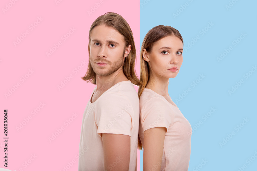 Powerful together. Charming young woman and her boyfriend standing back-to-back and looking at the camera while posing blue and pink backgrounds respectively Powerful together. Charming young woman and her boyfriend standing back-to-back and looking at the camera while posing blue and pink backgrounds respectively
