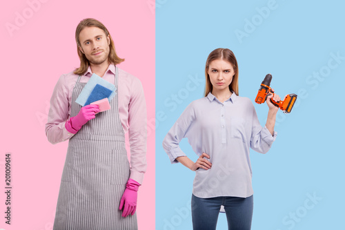 No gender stereotypes. Pretty young woman lifting up a screw gun and posing with it while her boyfriend holding sponges and wearing an apron