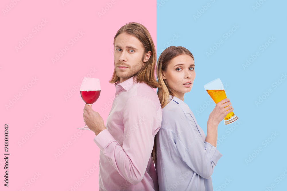 Enjoying alcohol. Pleasant young couple standing back-to-back and posing while the man holding a glass of wine and a woman having beer Enjoying alcohol. Pleasant young couple standing back-to-back and posing while the man holding a glass of wine and a woman having beer