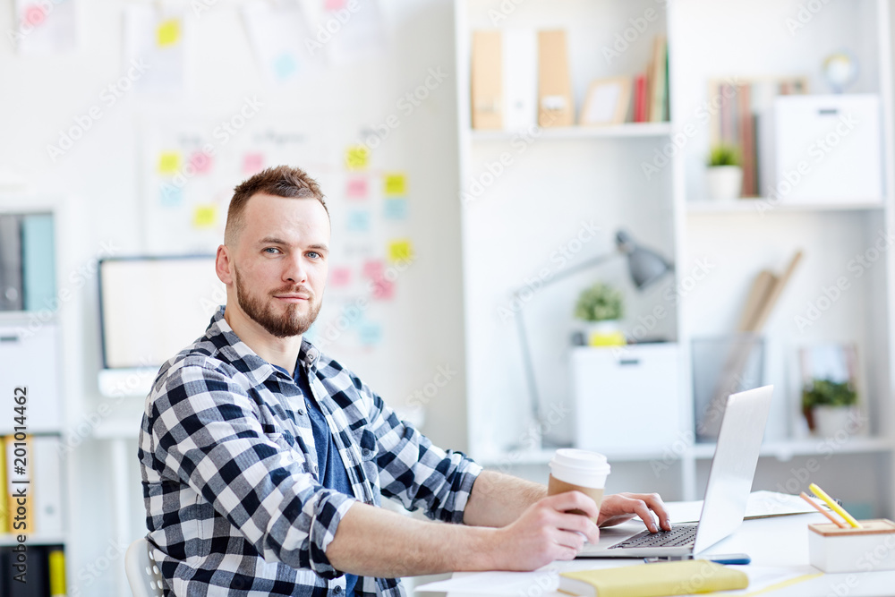 Portrait of young handsome bearded man in checked shirt looking at camera while sitting at laptop with coffee in modern office Portrait of young handsome bearded man in checked shirt looking at camera while sitting at laptop with coffee in modern office