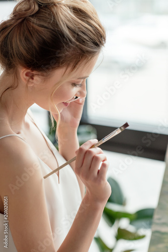 beautiful smiling young painter holding brush and talking by smartphone in art studio