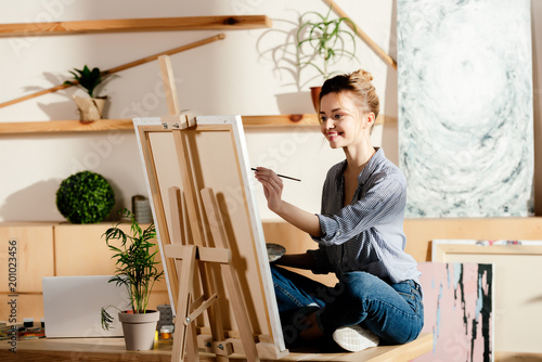 smiling female artist sitting on table and drawing on canvas in studio