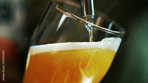 Process of pouring beer into glass beaker in grill bar. Alcoholic beverage is poured into transparent glass with help of portioning equipment indoors. Yellow foamed drink is flowing by thin stream