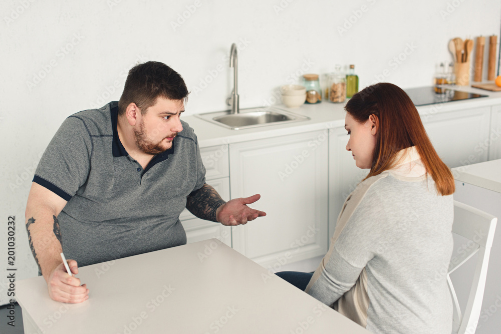 overweight boyfriend quarreling with girlfriend and holding smartphone in kitchen overweight boyfriend quarreling with girlfriend and holding smartphone in kitchen