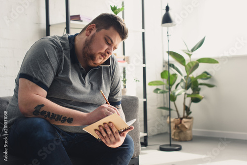 Handsome overweight man writing something to notebook on sofa at home photo