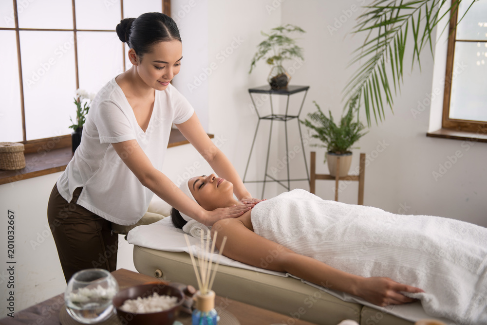 Massage session. Cheerful pleasant woman standing near her client while doing a relaxing massage for her