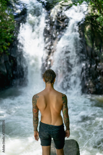 back view of tattooed man standing with Aling-Aling Waterfall on background, Bali, Indonesia