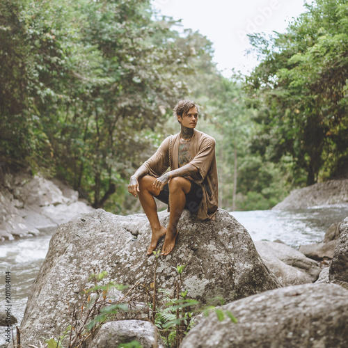 tattooed man resting on rock with green plants and river on backdrop  Bali  Indonesia