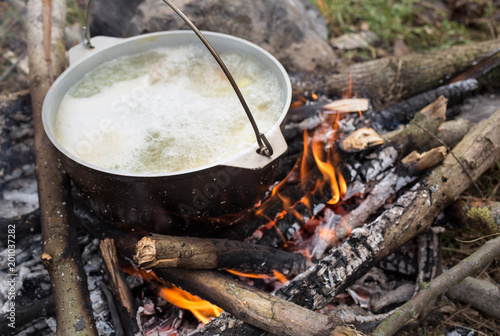 Old vintage pot in black soot is standing on the open fire in the wood. Natural summer holidays photo. Fish broth cooked in the forest. Vegetarian carrots and potatoes vegetable soup.