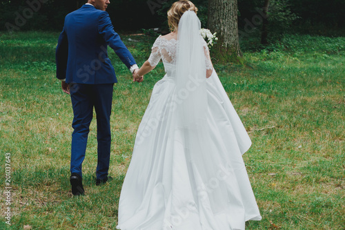 Wedding couple are holding hands in the green forest. Groom and bride in dress with train and tulle veil are walking outdoors. High green trees and bright emerald green grass.