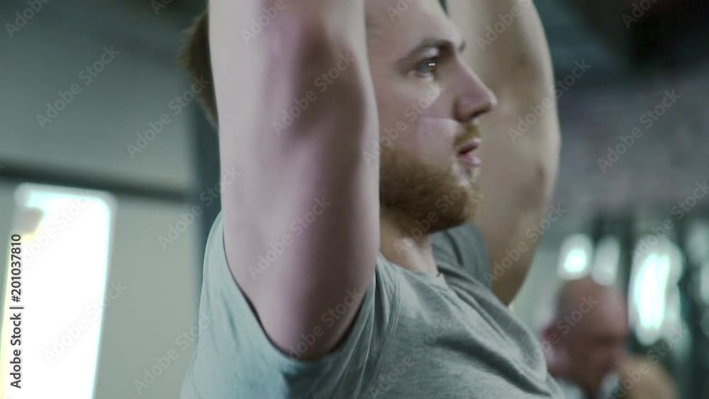 Portrait of young sportsman, who is doing barbell presses in modern gym. Handsome man in grey t-shirt is staning, holding weight equipment by his muscular hands and working on his biceps and Portrait of young sportsman, who is doing barbell presses in modern gym. Handsome man in grey t-shirt is staning, holding weight equipment by his muscular hands and working on his biceps and