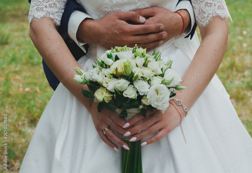 Wedding couple is hugging in the green forest. Bride in satin lace dress with train and tulle veil is holding a bouquet. Bright emerald green grass outdoors. White roses and eustoma flowers. Wedding couple is hugging in the green forest. Bride in satin lace dress with train and tulle veil is holding a bouquet. Bright emerald green grass outdoors. White roses and eustoma flowers.