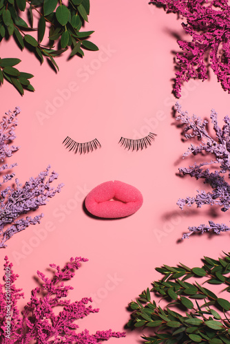 top view of face made of sponge and false eyelashes surrounded with flowers on pink surface