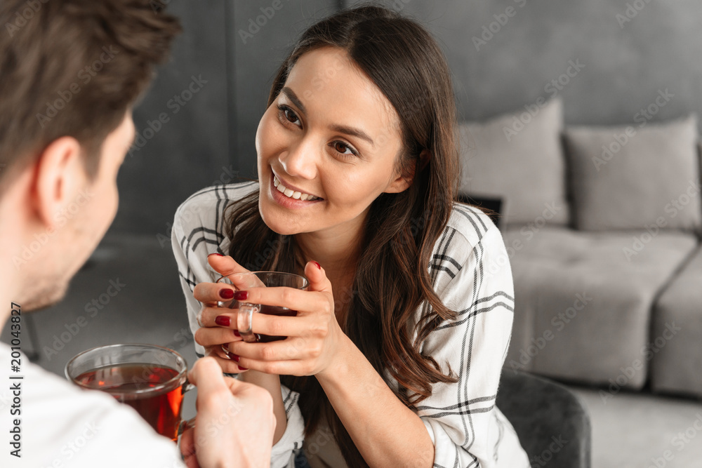 Photo of beautiful smiling woman looking at man with love, while sitting at table in flat and drinking tea from glass Photo of beautiful smiling woman looking at man with love, while sitting at table in flat and drinking tea from glass
