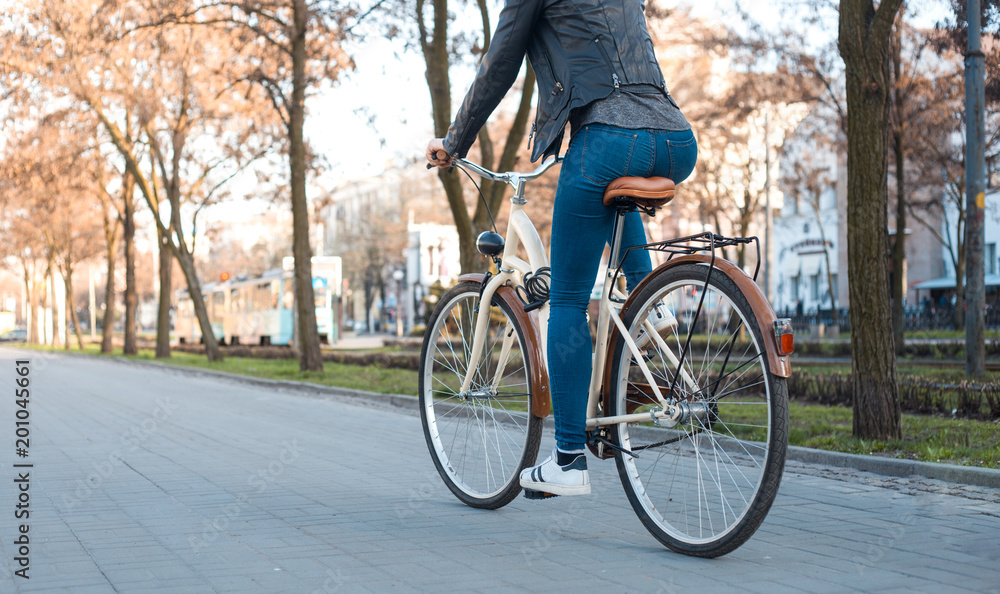 Fototapeta premium A girl in a leather jacket and jeans rides a bicycle along the avenue in the spring