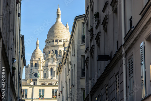 View of the facade and dome of the Basilica of the Sacred Heart of Paris through a narrow street between old parisian buildings under a clear blue sky. © olrat