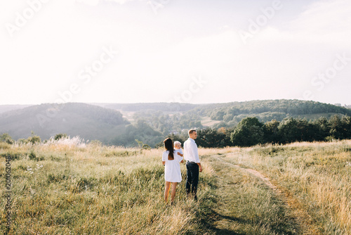 Walk beautiful young family in white clothes with a young son blond in mountainous areas with tall grass at sunset. Mother keeps son in her arms  hugging. family - this is happiness