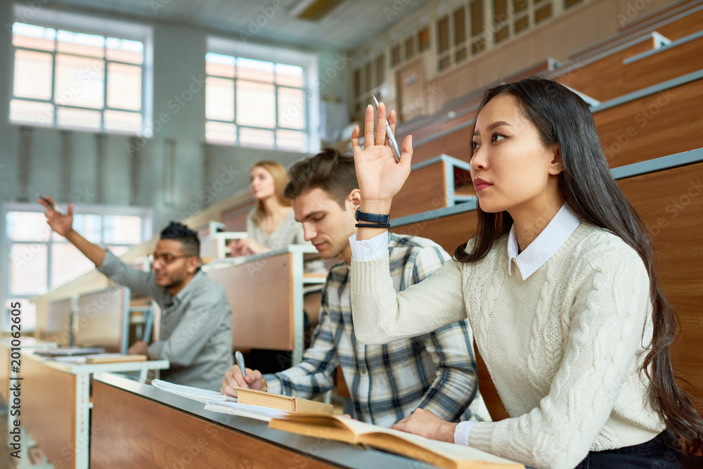 © Seventyfour - Multi-ethnic group of students sitting in row at tables in lecture hall of modern college and raising hands, focus on pretty Asian woman in foreground