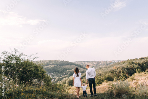 Walk beautiful young family in white clothes with a young son blond in mountainous areas with tall grass at sunset. Mother keeps son in her arms, hugging. family - this is happiness