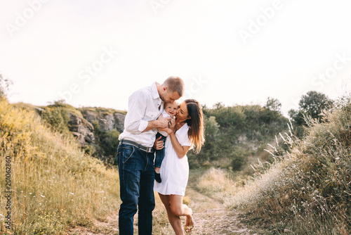 Walk of beautiful young family in white clothes with a young son blond in mountainous areas with tall grass at sunset. Dads carries his son on his shoulders. family - this is happiness © Oleksandr