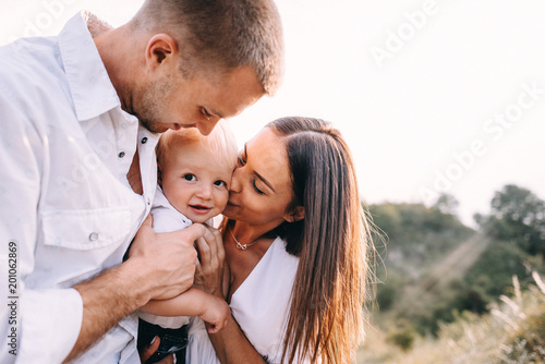 Walk of beautiful young family in white clothes with a young son blond in mountainous areas with tall grass at sunset. Dads carries his son on his shoulders. family - this is happiness