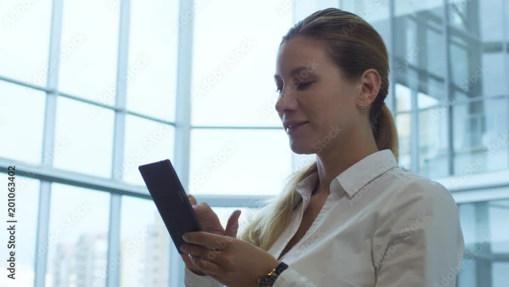 Panning shot of mid-aged attractive business lady standing in office center and typing on smart phone