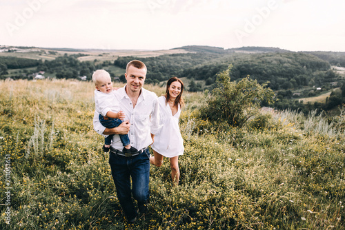 Walk beautiful young family in white clothes with a young son blond in mountainous areas with tall grass at sunset. Mother keeps son in his arms  hugging. family - this is happiness