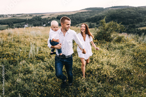 Walk beautiful young family in white clothes with a young son blond in mountainous areas with tall grass at sunset. Mother keeps son in his arms, hugging. family - this is happiness