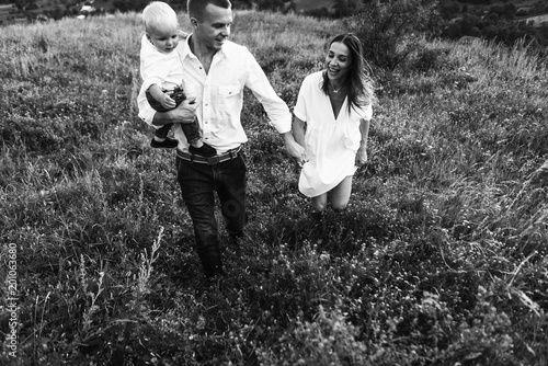 Walk beautiful young family in white clothes with a young son blond in mountainous areas with tall grass at sunset. Mother keeps son in his arms, hugging. family - this is happiness