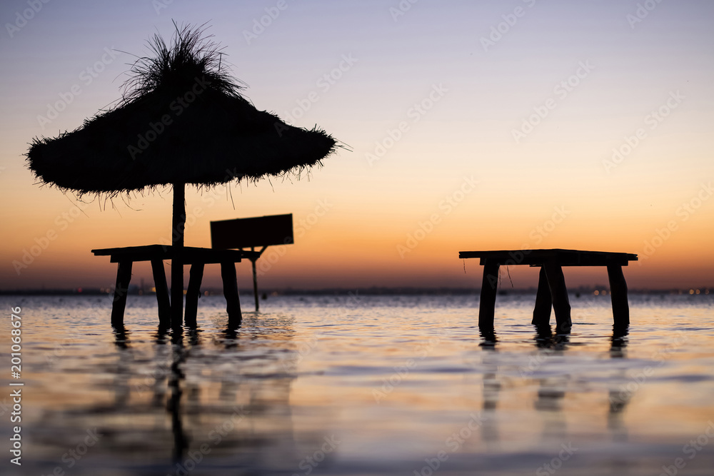 Black silhouette of Exotic beach buildings - tables and a canopy standing on the beach in the water for tourists to relax at luxury vacation at sunset among the blue waves in Mediterranean sea Black silhouette of Exotic beach buildings - tables and a canopy standing on the beach in the water for tourists to relax at luxury vacation at sunset among the blue waves in Mediterranean sea