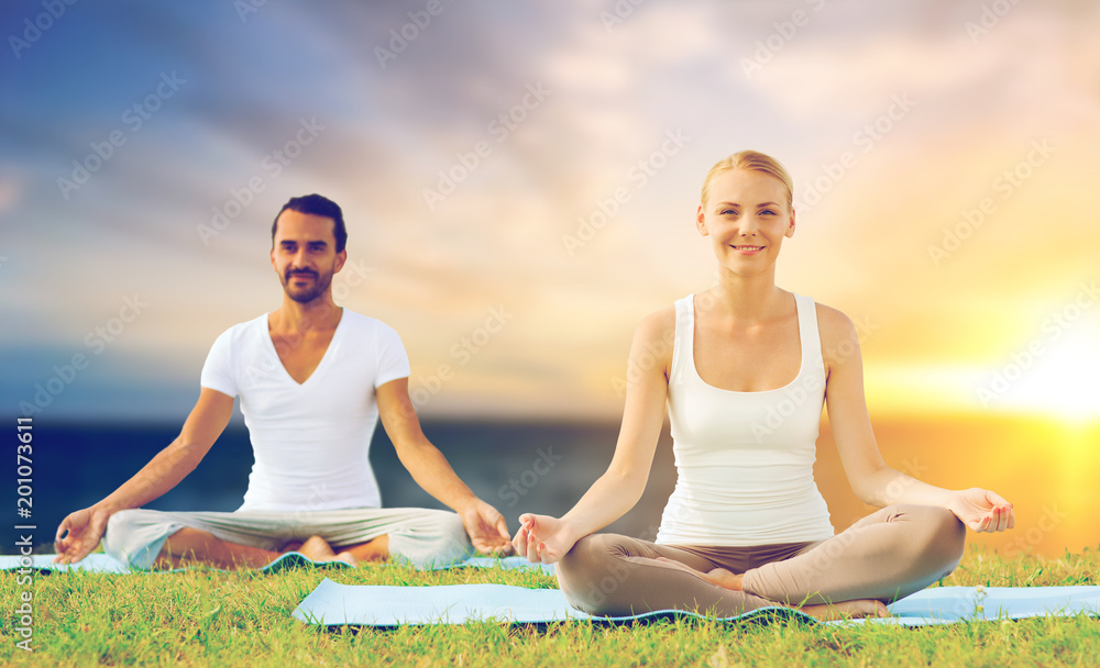 yoga , mindfulness, harmony and people concept - happy couple meditating in lotus pose outdoors over sea background