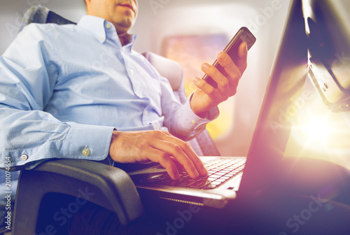 transport, tourism and technology concept - close up of businessman with smartphone and laptop traveling by plane and working over porthole background