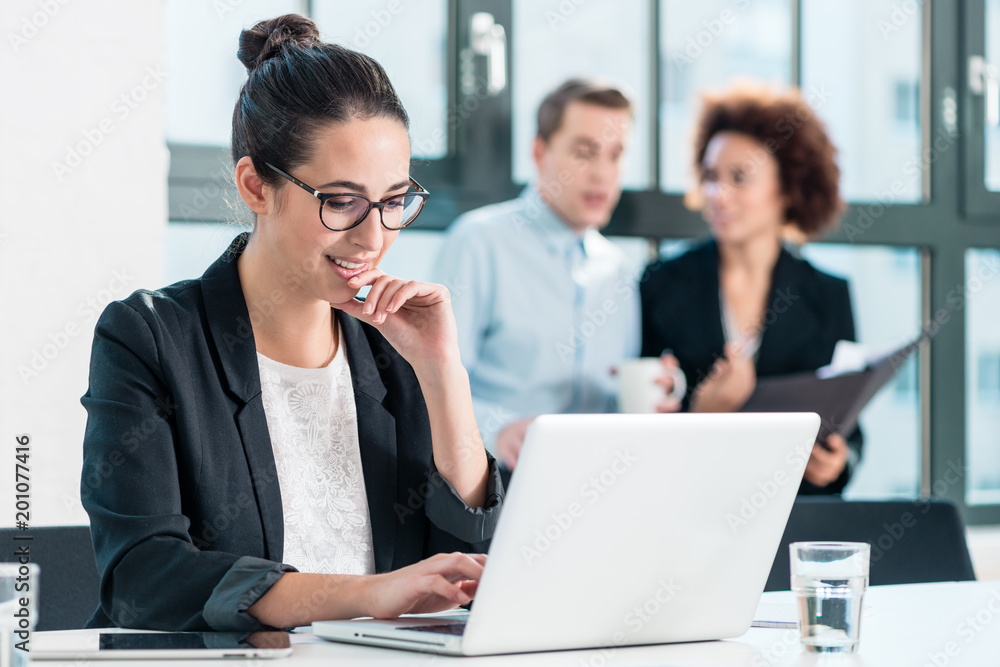 Young woman smiling while using a laptop at desk in the office in front of her colleagues Young woman smiling while using a laptop at desk in the office in front of her colleagues