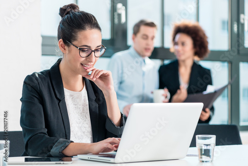 Young woman smiling while using a laptop at desk in the office in front of her colleagues 