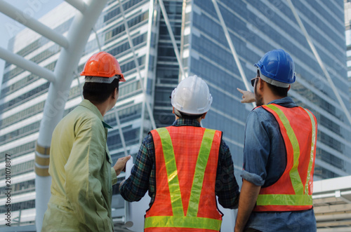 back view group of engineer, technician and architect with safety helmet planning about building plan with blueprint in modern city building background, construction site, industry and worker concept
