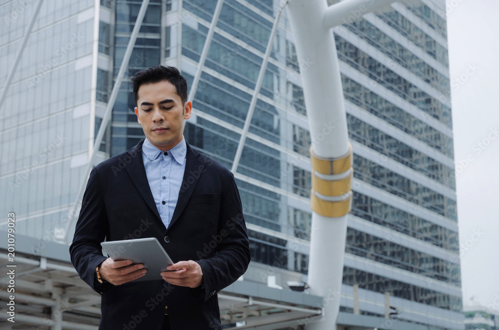 young handsome asian business man wearing modern black suit reading information about finance news with mobile tablet in building city background, network technology, internet, successful concept