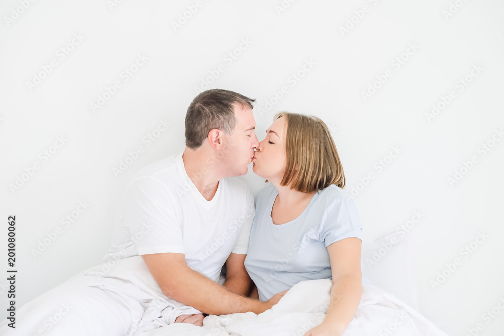 Kissing cute couple in sleepwear on the bed. Husband holding hand on stomach of his pregnant wife. Happy and loving family morning concept. Soft selective focus. Space for text. Kissing cute couple in sleepwear on the bed. Husband holding hand on stomach of his pregnant wife. Happy and loving family morning concept. Soft selective focus. Space for text.