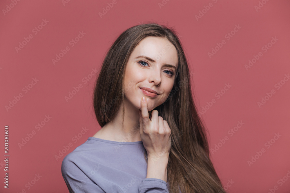 Pleased attractive female with appealing appearance, keeps hand under chin, has luxurious brunette hair, looks delightfully at camera, isolated over pink background. Positive emotions concept