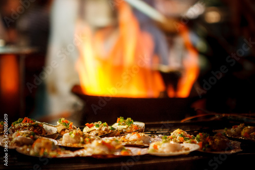 Grilled scallops sold at a street market stall with a pan covered with flames on the background. Chinatown, Bangkok, Thailand