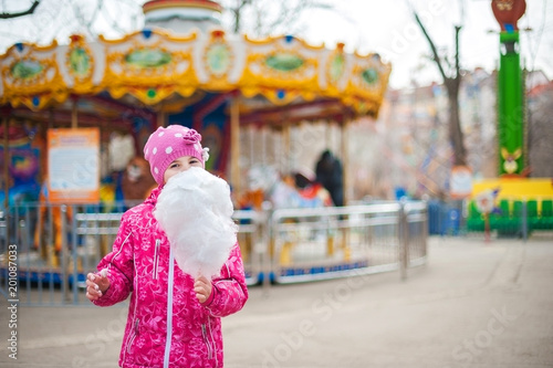 A photograph of a child's rest on nature in the spring. A child girl in a bright pink jacket is eating sweet cotton wool against the backdrop of rides, carousels. Close-up portrait with sweet cotton w
