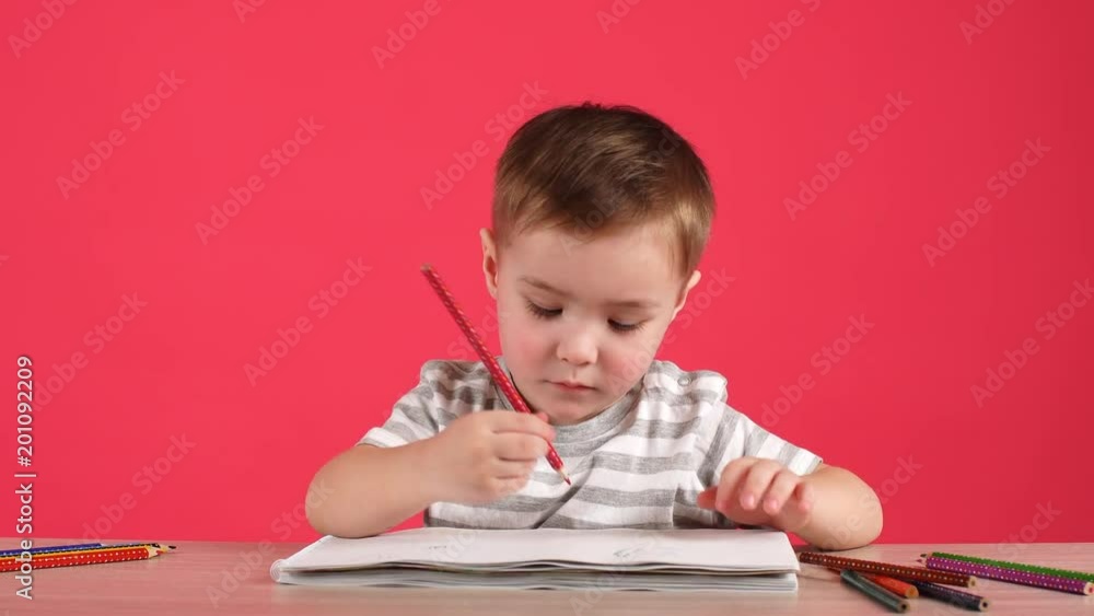 Cute baby boy drawing with pencils in album on pink background. Cute baby boy drawing with pencils in album on pink background.