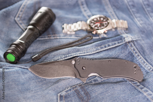 Folding knife, searchlight and men's watch on the jeans backgound
