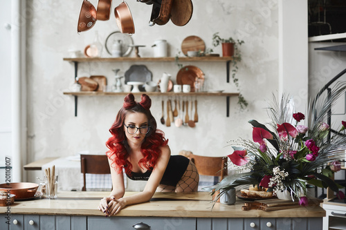 Beautiful and sexy pin-up model girl with red hair and professional makeup, in black bodysuits and glasses posing on a kitchen