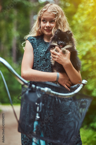 Portrait of a little blonde girl in a casual dress, holds cute spitz dog, in a park.