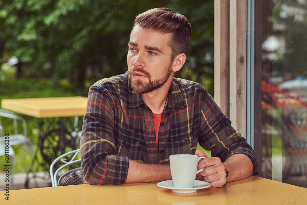 A handsome fashionable male with stylish haircut and beard, wearing fleece shirt, drinking coffee at the cafeteria. A handsome fashionable male with stylish haircut and beard, wearing fleece shirt, drinking coffee at the cafeteria.