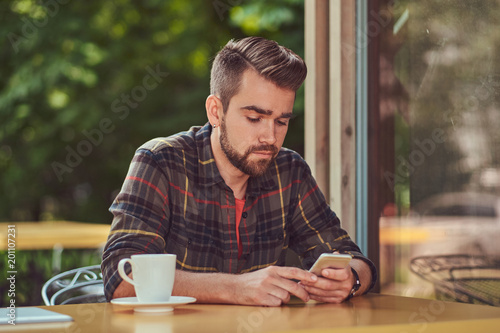 A handsome fashionable male with stylish haircut and beard, wearing fleece shirt, drinking coffee and holding a smartphone in the cafeteria.